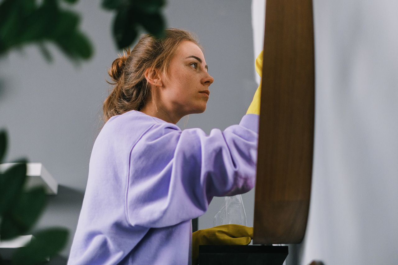 about-us-02 Vue de côté d’une jeune femme concentrée, portant des gants en latex, debout et nettoyant un miroir dans une pièce lumineuse en journée